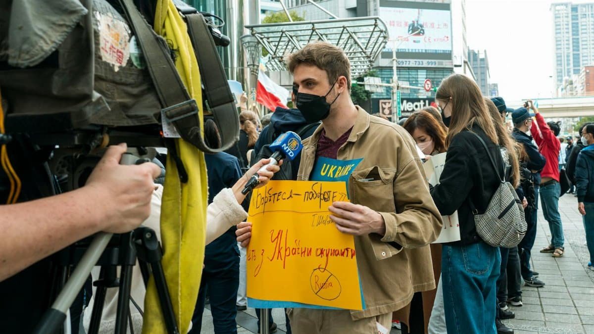 Young man holds protest sign during street demonstration against war, covered by media.