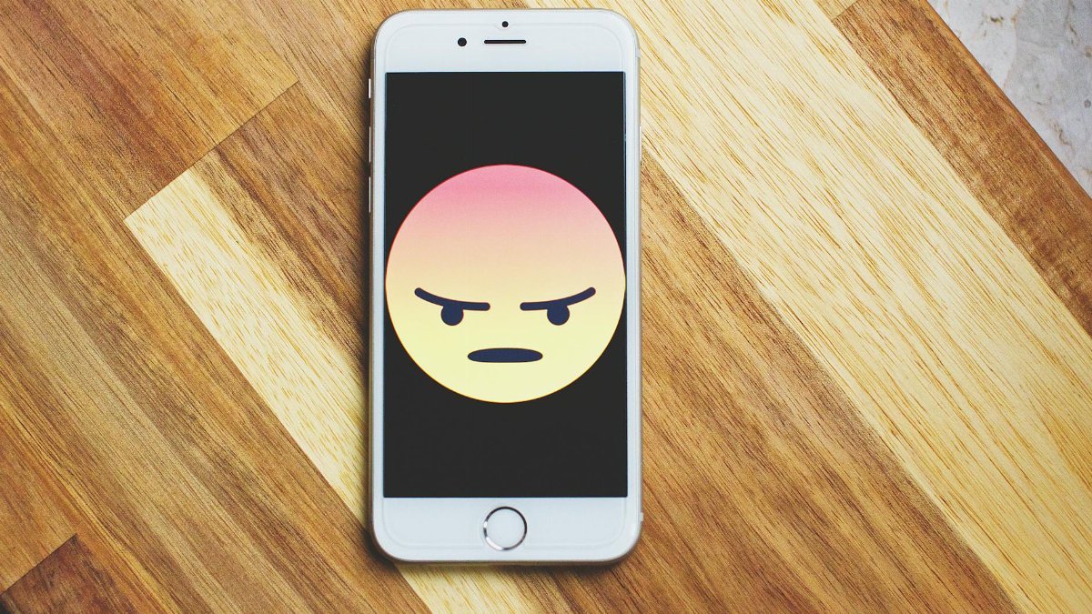 A smartphone showing an angry emoticon rests on a wooden table, emphasizing tech and emotion.