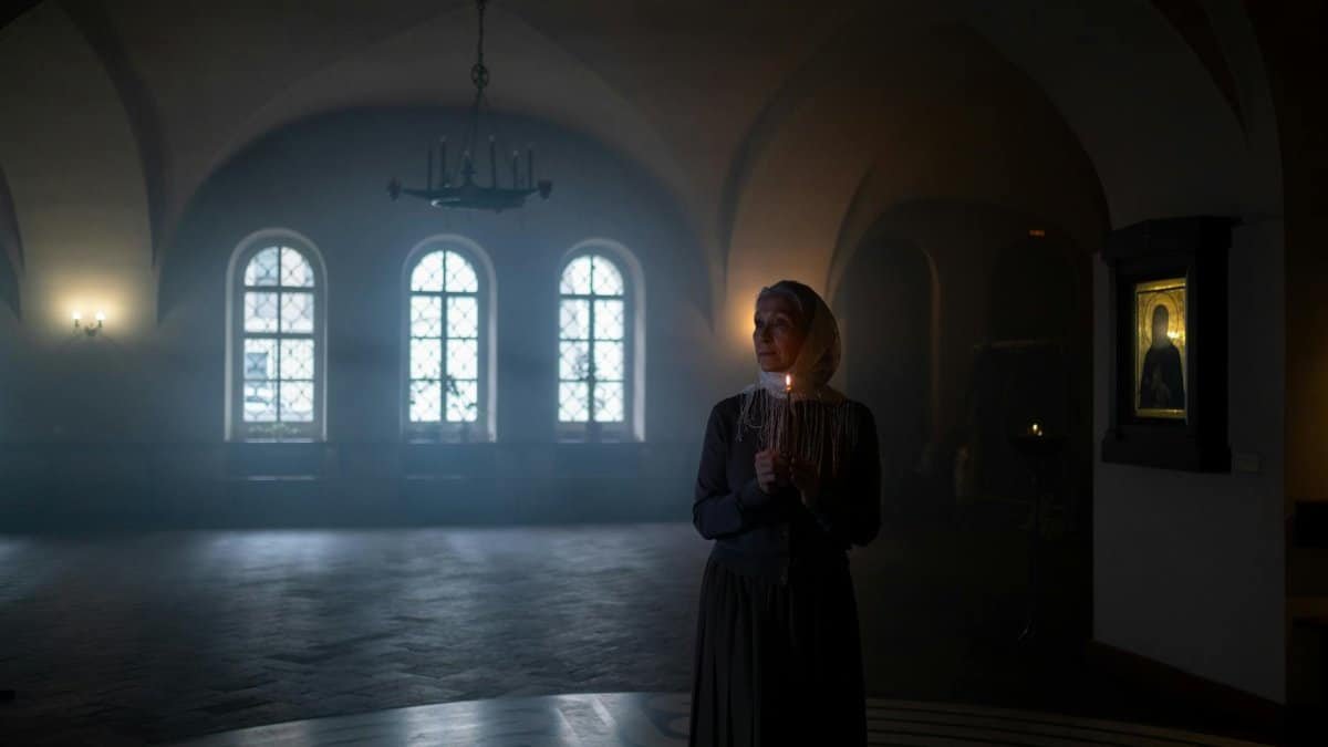 An elderly woman holds a candle in a serene, dimly lit church hallway, creating a spiritual atmosphere.
