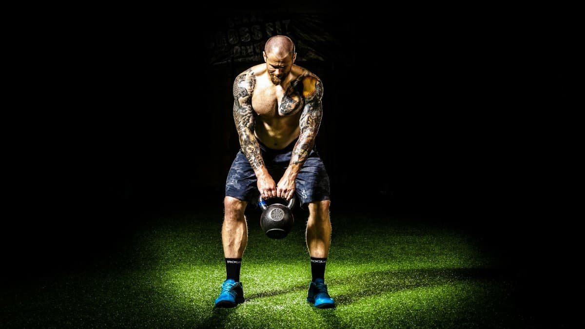 Muscular man with tattoos lifting a kettlebell indoors under a spotlight. Intense fitness and strength training.