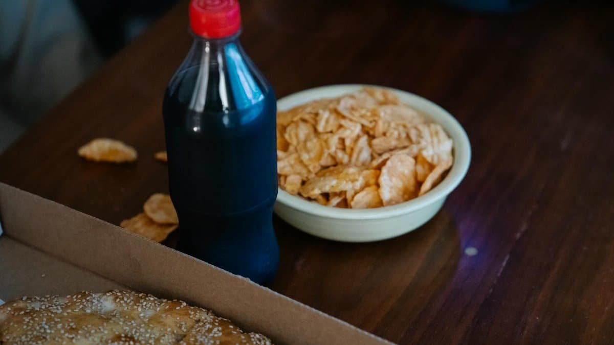 From above of plastic bottle of soft drink between takeaway pizza with sesame seeds and crispy potato chips at home