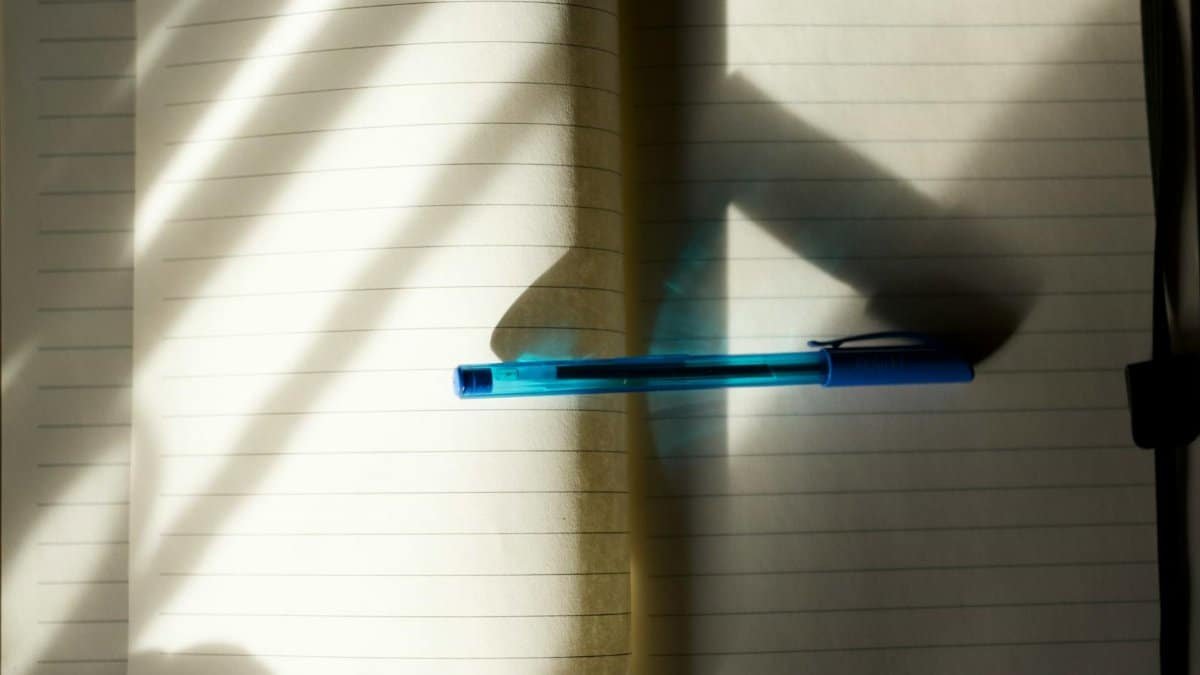 An open notebook with a blue pen in natural sunlight, casting shadows.