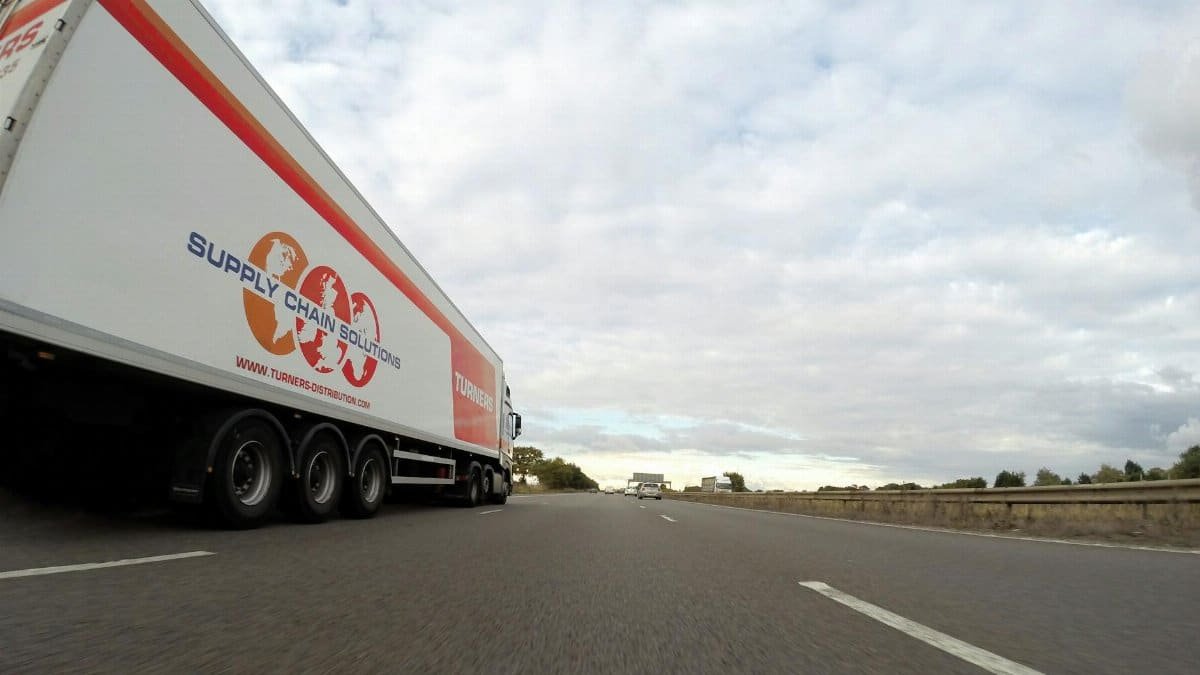 A large semi truck labeled 'Supply Chain Solutions' travels down a highway under a cloudy sky.
