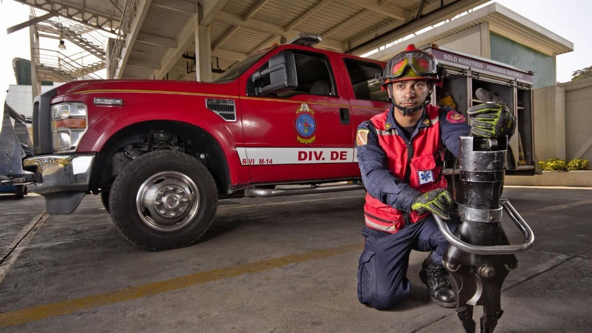 Firefighter in safety gear kneeling by a firetruck holding hydraulic rescue tool, showcasing bravery and readiness.