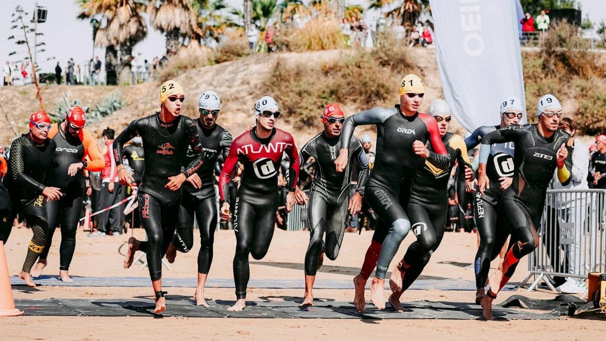 Full body of faceless slim barefoot sportspeople in wetsuits and swim caps running on sandy beach next to palms on slope in daytime
