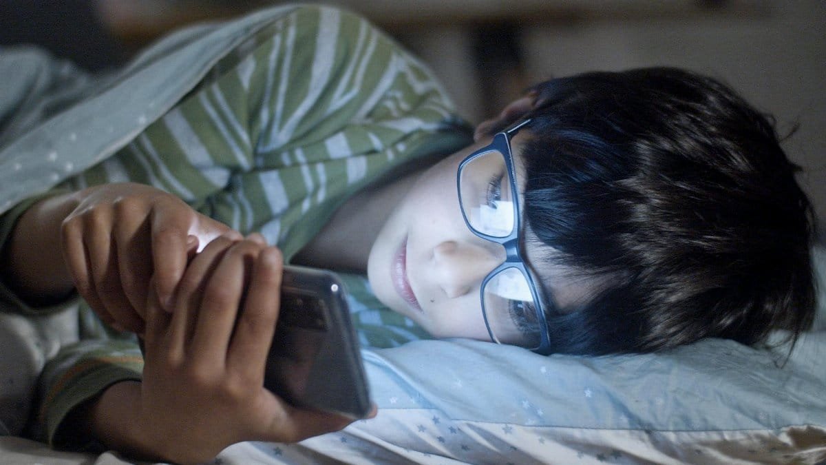 A child lying in bed using a smartphone at night, wearing glasses.