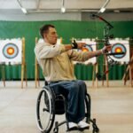 Focused young man in a wheelchair practicing archery indoors, aiming with determination at a target.