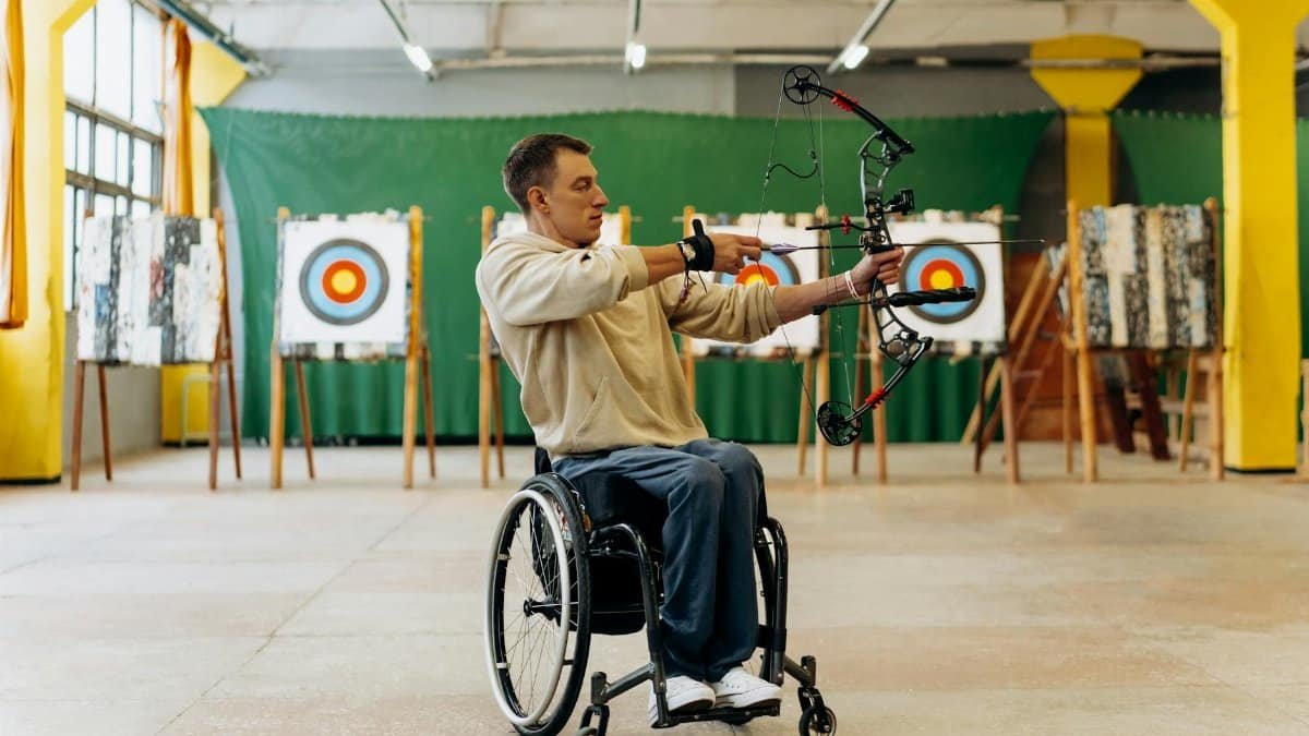 Focused young man in a wheelchair practicing archery indoors, aiming with determination at a target.