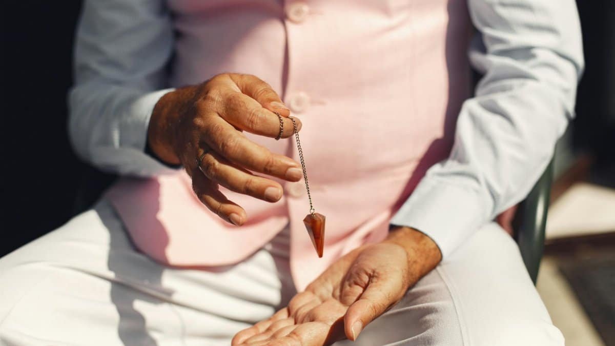 A man in soft pastel attire holding a gemstone pendulum over his palm outdoors.