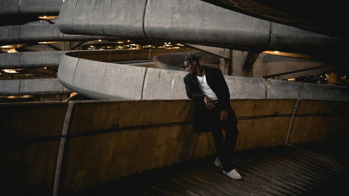 Full body of young stylish African American male in casual clothes looking out balcony of multistory parking garage against urban background