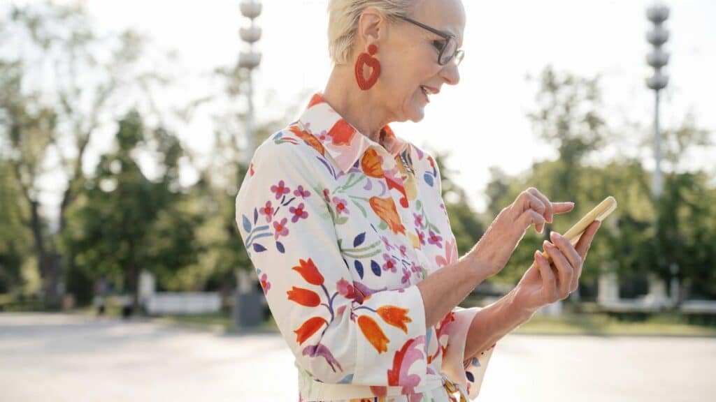 Smiling senior woman in floral dress using smartphone outdoors on sunny day.