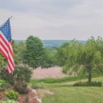 A serene suburban house porch displaying an American flag with a scenic background.