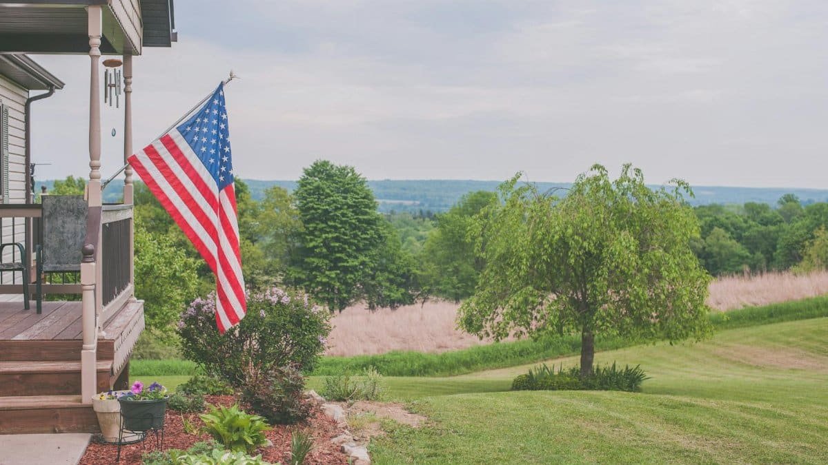 A serene suburban house porch displaying an American flag with a scenic background.