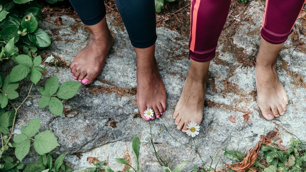 Two people standing barefoot on rocky ground with daisies on toes outdoors.