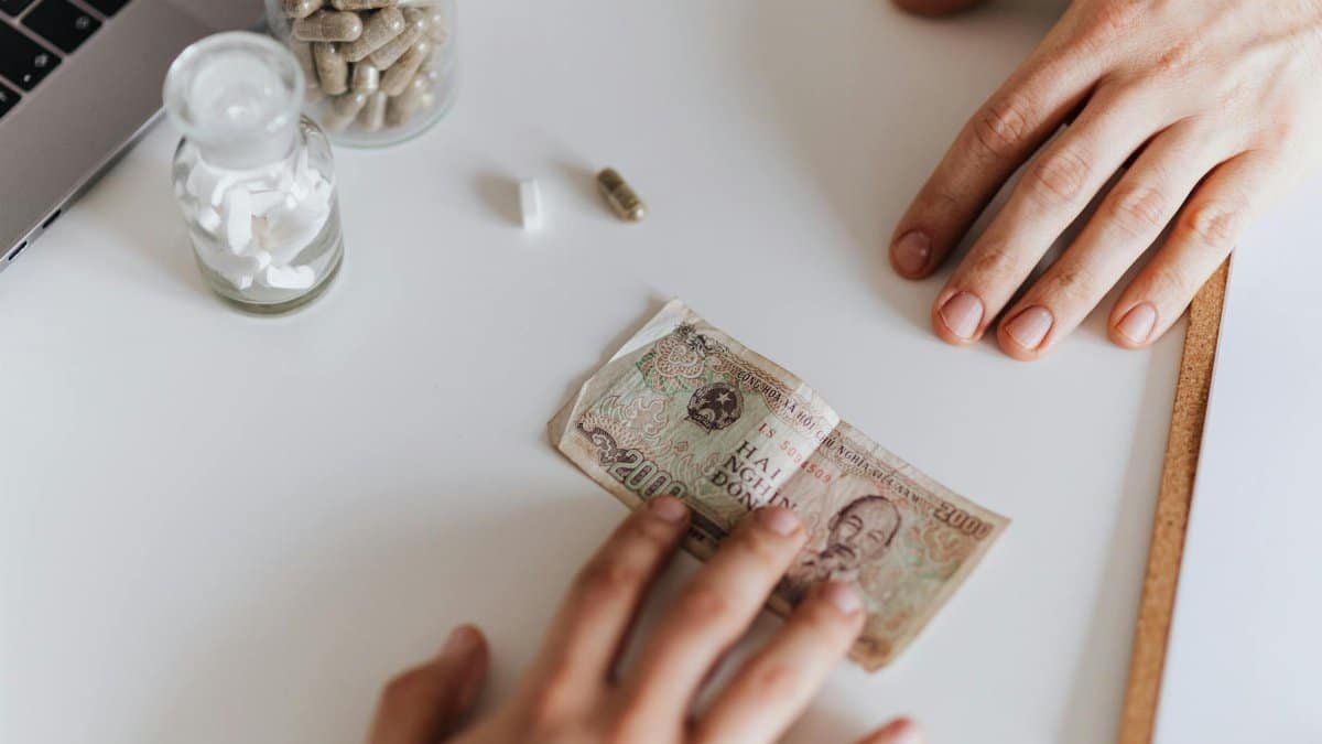 Conceptual photo showing hands exchanging medicine and currency on a desk.
