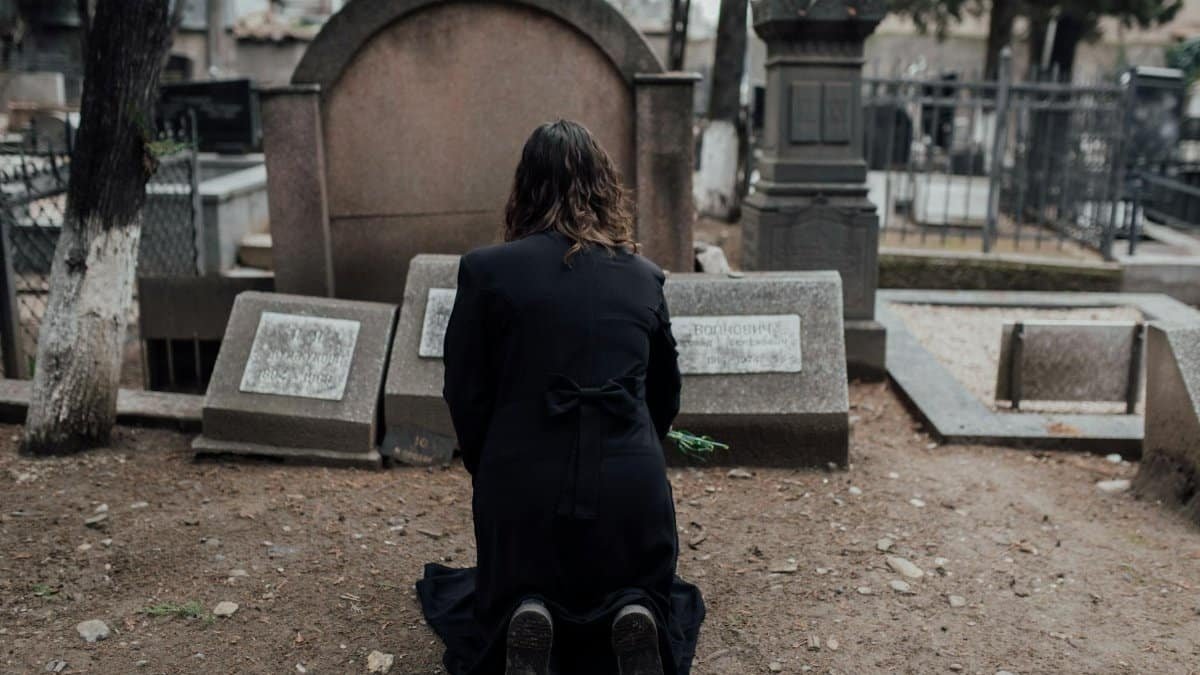 A woman in black kneels at a memorial in a quiet cemetery setting.