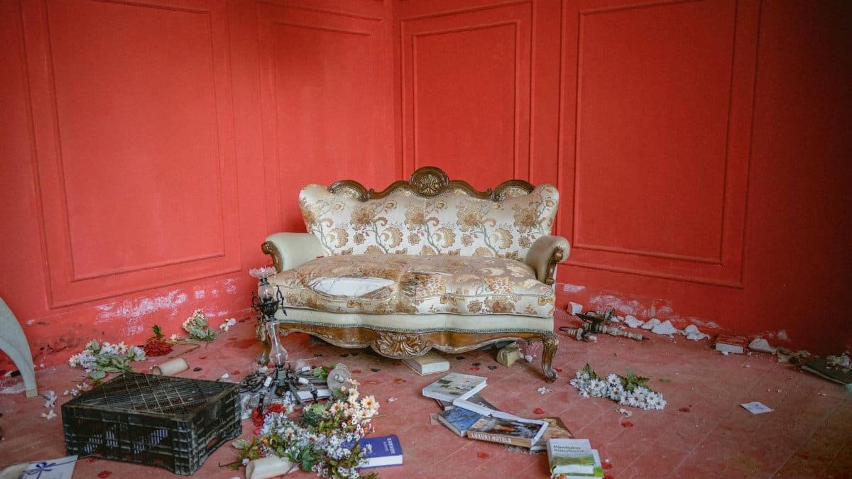 A vintage sofa sits in a neglected room with red walls, scattered books, and flowers.