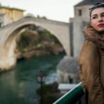 Fashionable woman in fur coat posing near a historic stone bridge during the day.