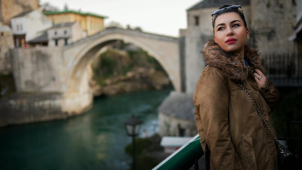 Fashionable woman in fur coat posing near a historic stone bridge during the day.