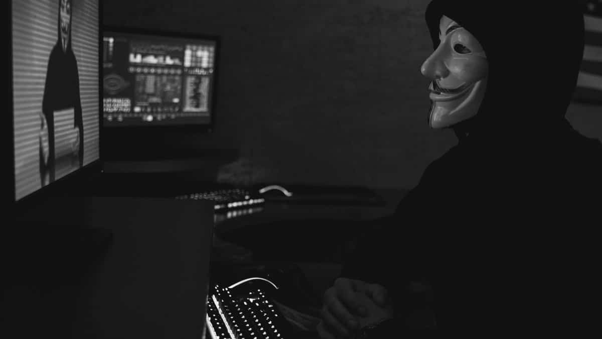 Monochrome image of a hacker in a Guy Fawkes mask working on a computer in a dimly lit room.