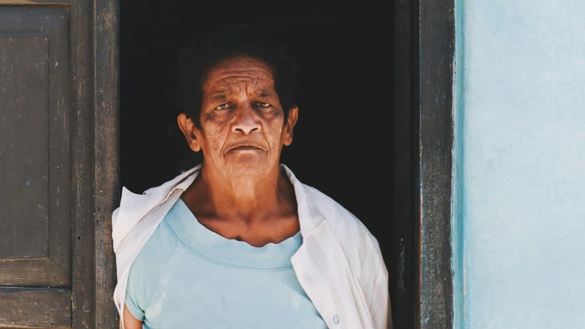 A senior woman in a blue blouse stands solemnly in a doorframe, conveying wisdom and experience.