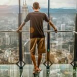 A man stands barefoot on a glass deck high above Kuala Lumpur, enjoying the stunning cityscape view.
