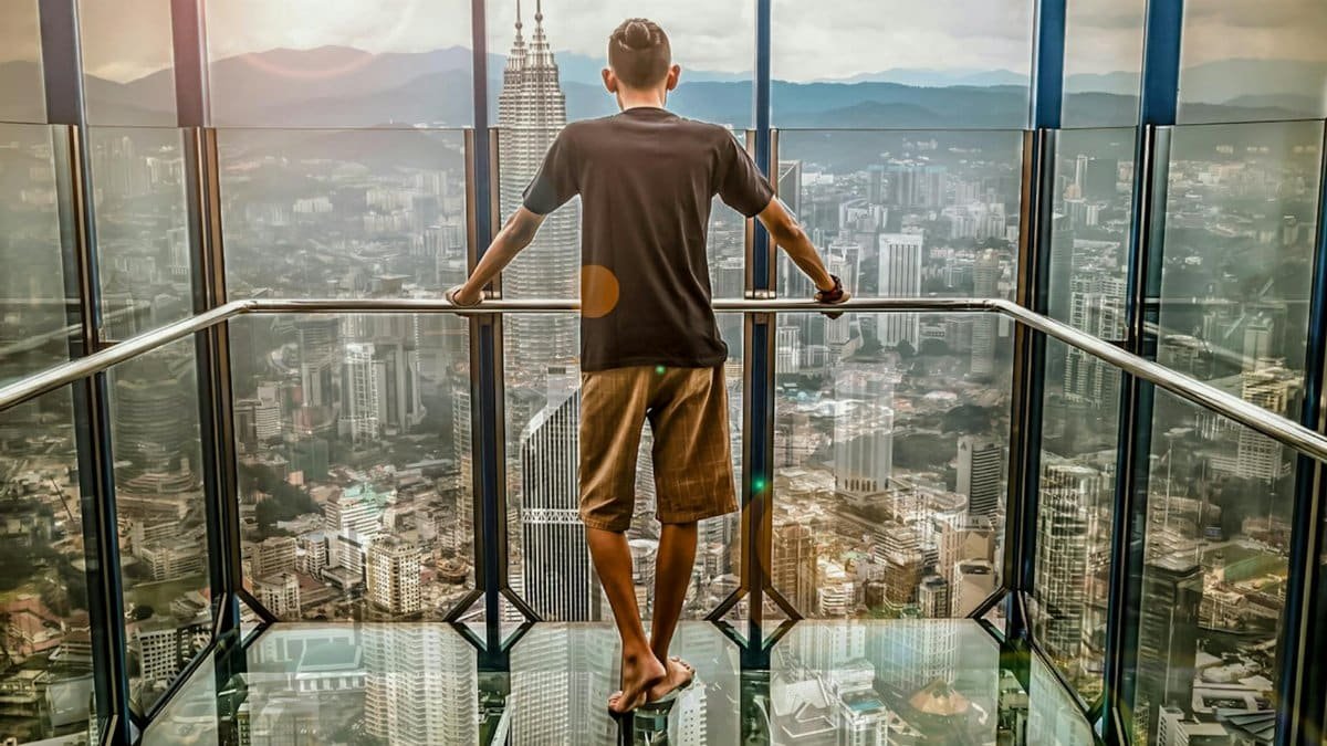 A man stands barefoot on a glass deck high above Kuala Lumpur, enjoying the stunning cityscape view.