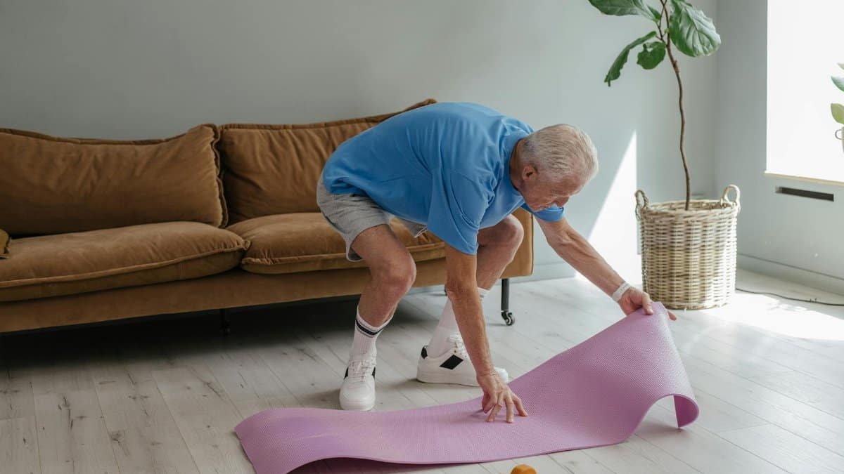 An elderly man sets up his yoga mat indoors, embodying a healthy lifestyle.