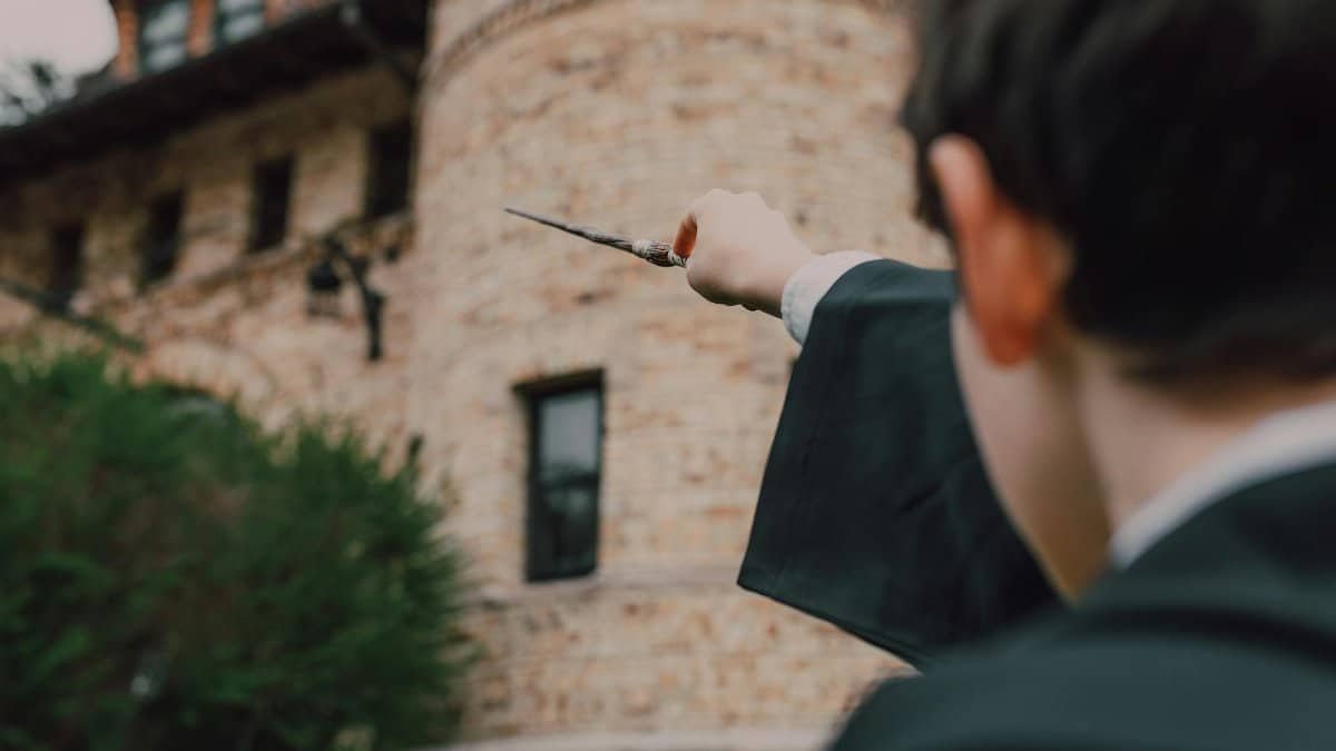 Child in wizard costume waving a wand outside a stone building, evoking magical themes.