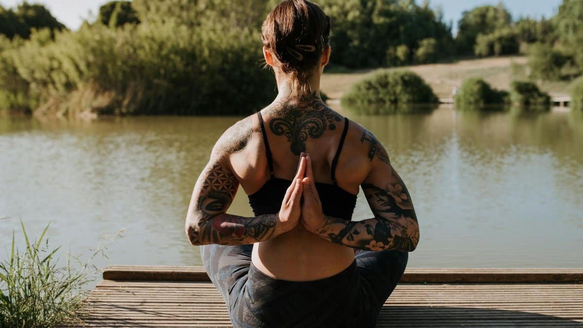 Back view of anonymous tattooed female in sportswear sitting in Reverse Prayer pose on wooden dock against river