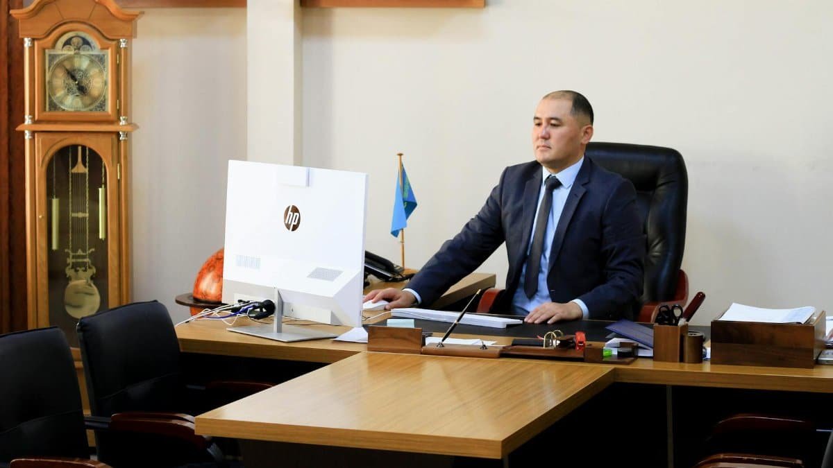 A serious businessman in a black suit focuses on work at his office desk with a computer and office decor.