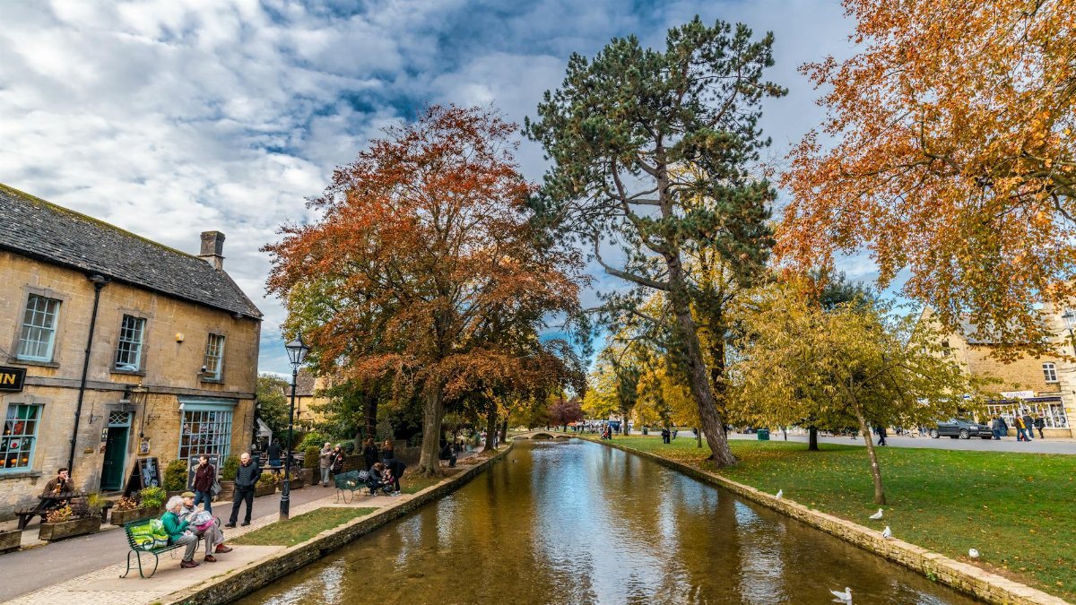 Charming autumn view of Bourton-on-the-Water with a tranquil stream and vibrant foliage.