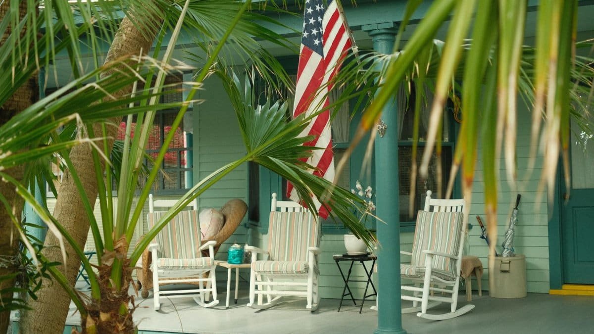 Charming Southern porch with American flag and rocking chairs, hidden behind lush palm trees.