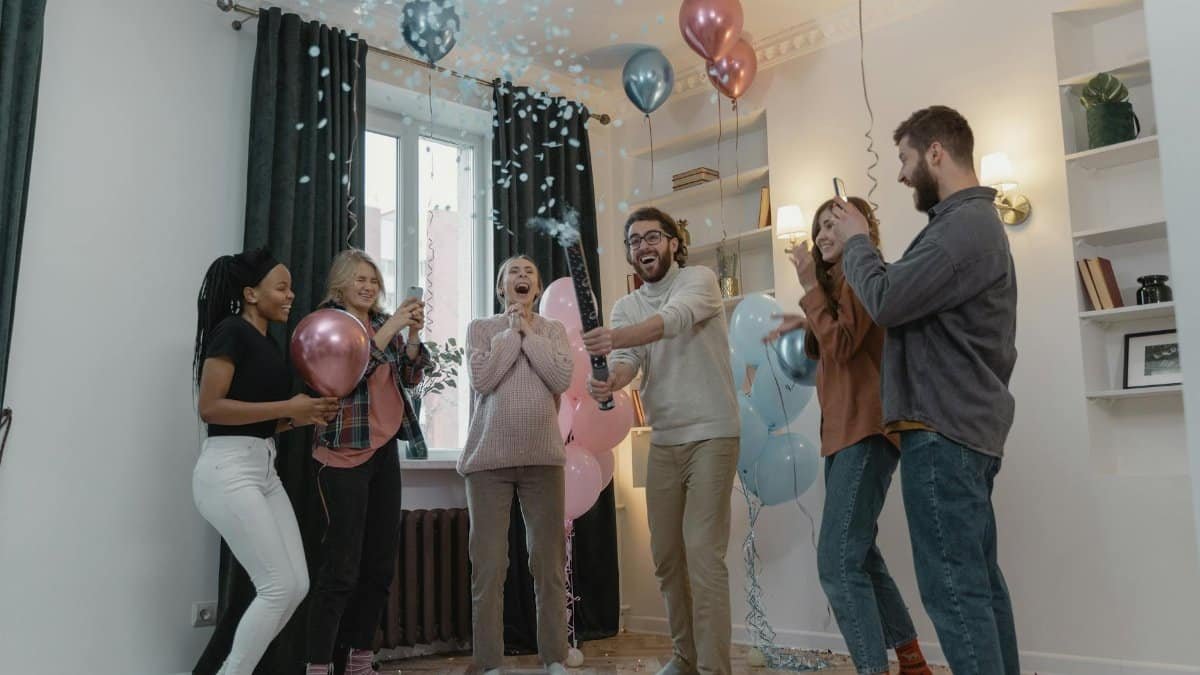 Excited friends celebrate a gender reveal with balloons and confetti indoors.