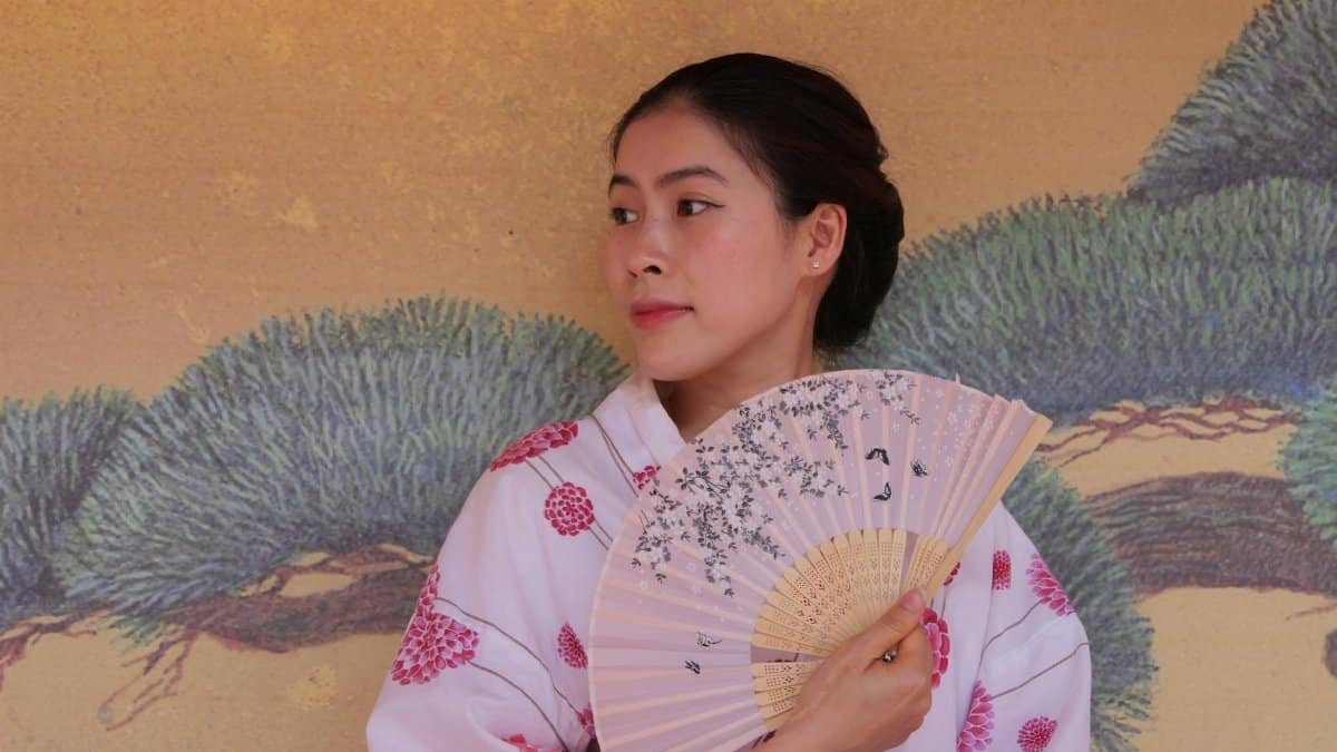 Asian woman holding a decorated fan, wearing a kimono in an artistic indoor setting.