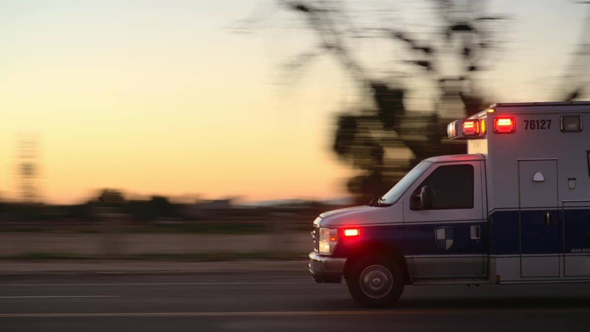 A fast-moving ambulance races down a city street at dusk, lights flashing urgently.
