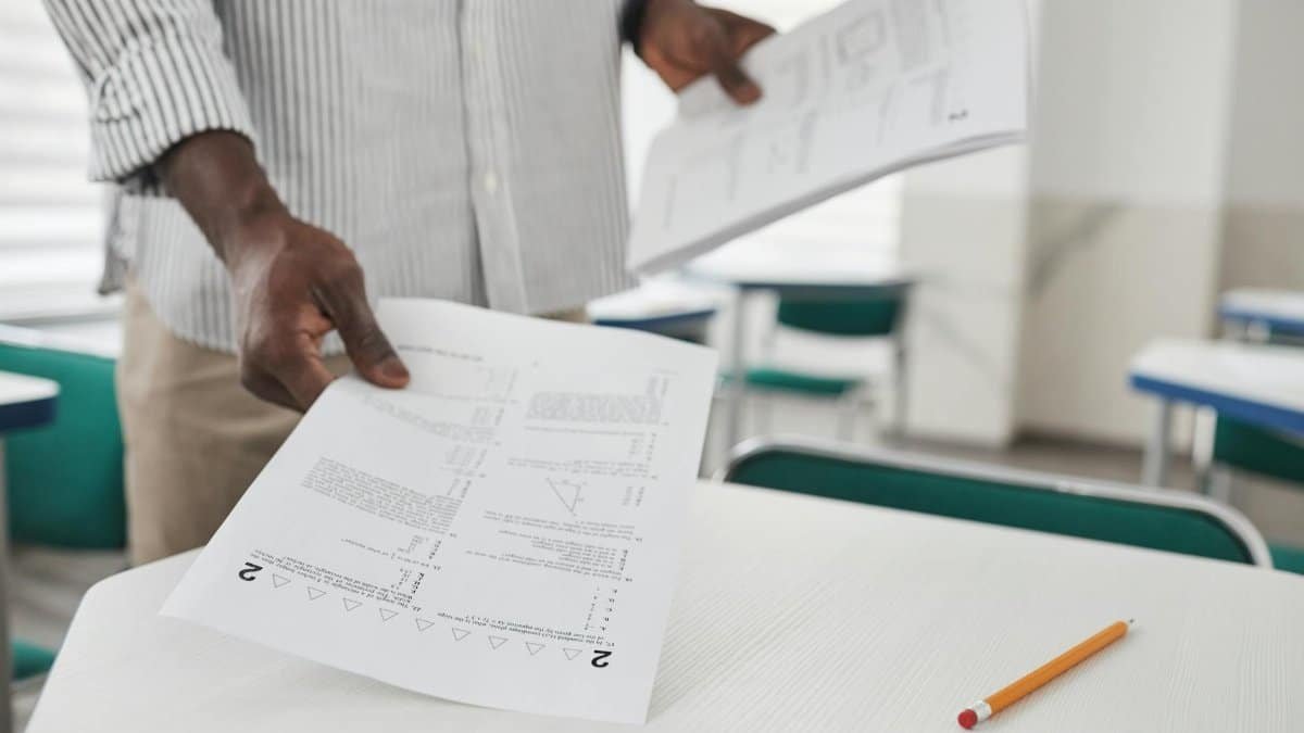 Close-up of a person arranging examination papers on a desk in a classroom.