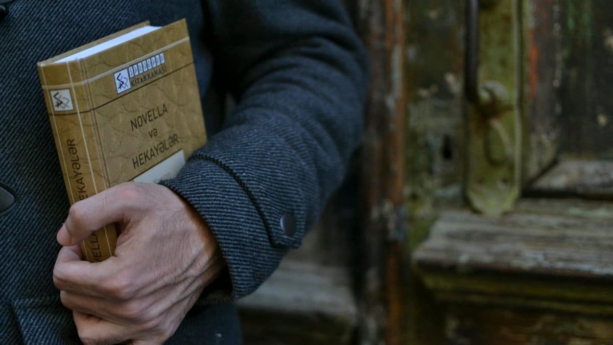 A hand holding a book titled 'Novella və Hekayələr' in front of a weathered wooden door.