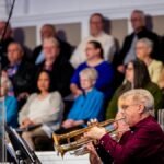 A trumpet player performs at an indoor concert surrounded by an audience, creating a lively musical atmosphere.