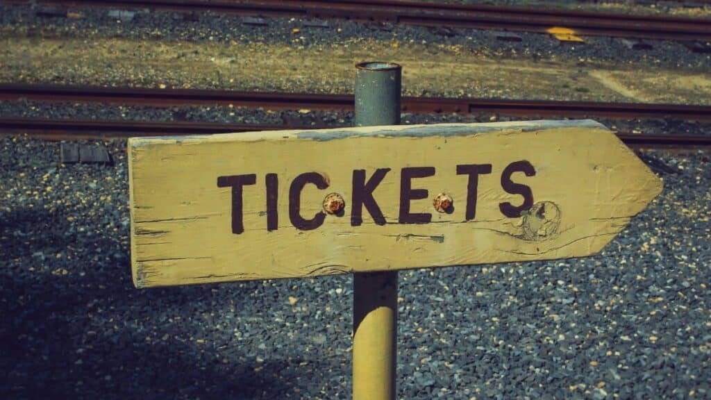 Weathered wooden ticket sign pointing towards railway tracks outdoors, vintage look.