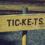 Weathered wooden ticket sign pointing towards railway tracks outdoors, vintage look.