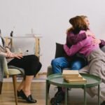 Side view of positive female therapist looking at loving couple cuddling on sofa near table with books
