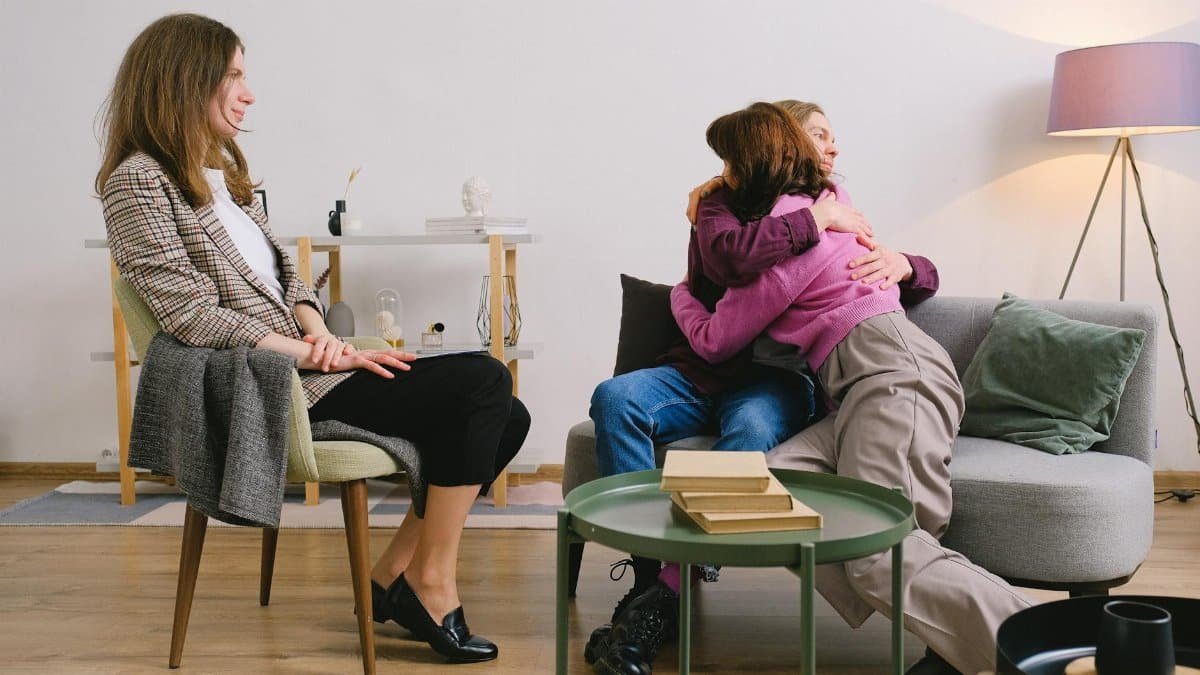 Side view of positive female therapist looking at loving couple cuddling on sofa near table with books
