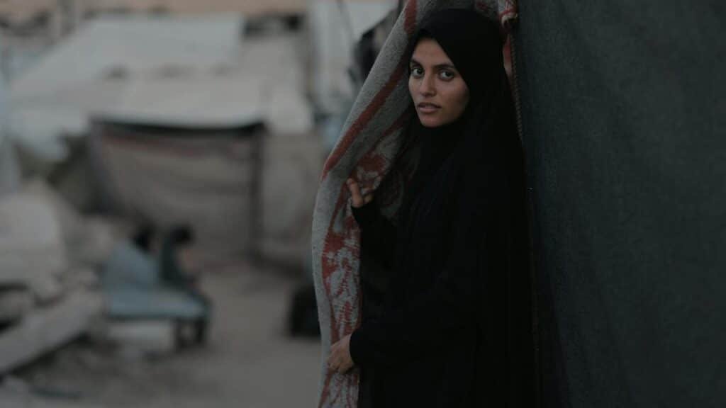 Portrait of a young woman standing in a Gaza refugee camp amidst rubble and tents.