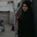 Portrait of a young woman standing in a Gaza refugee camp amidst rubble and tents.
