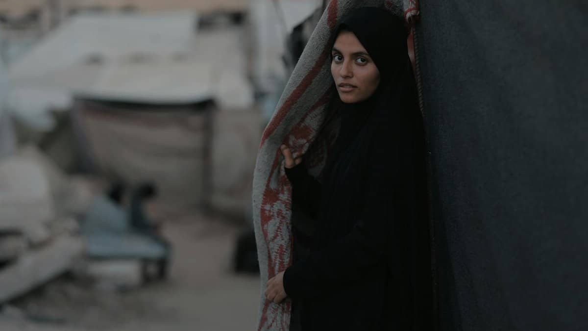 Portrait of a young woman standing in a Gaza refugee camp amidst rubble and tents.