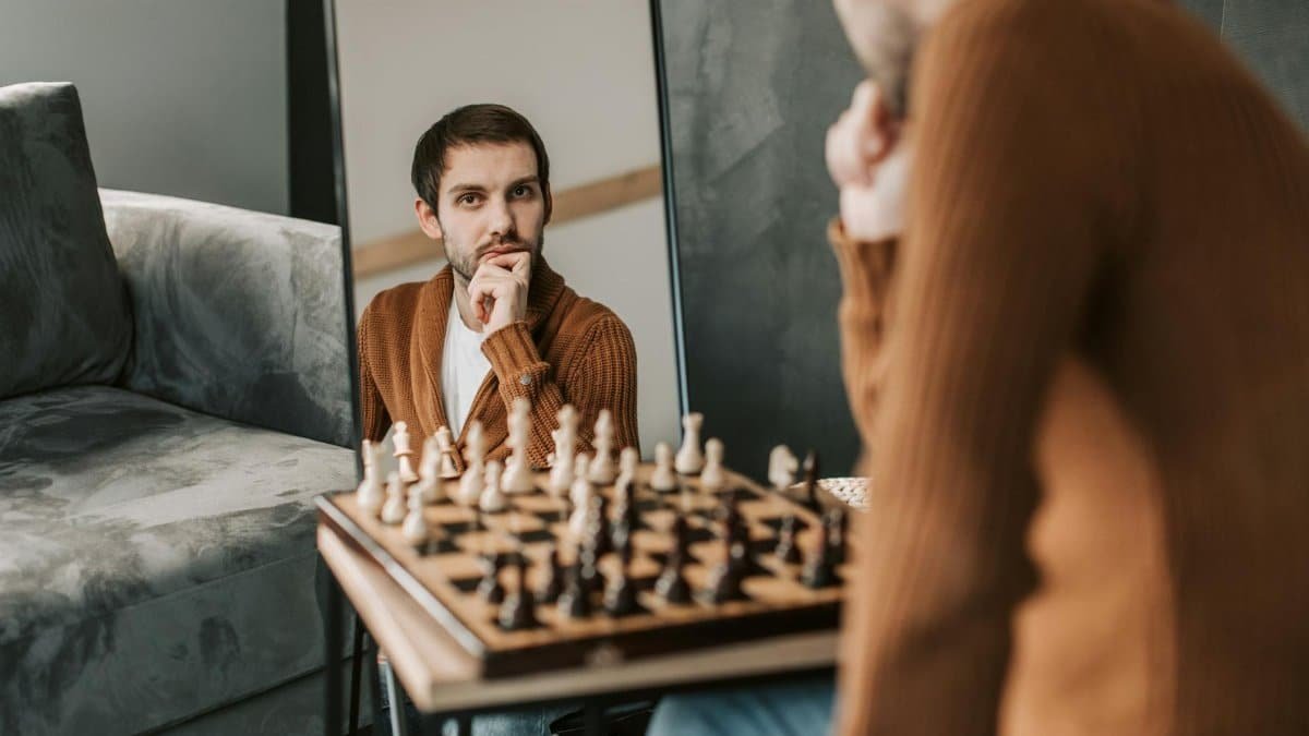 Man in brown sweater engages in chess, captured in thoughtful reflection through a mirror indoors.