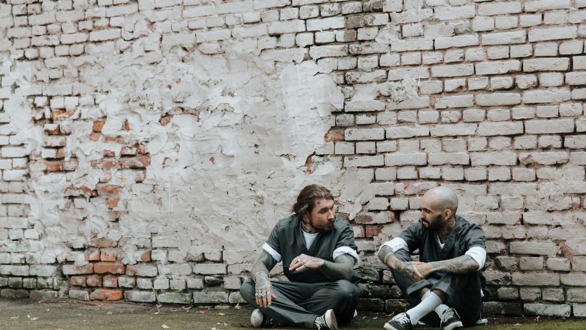Tattooed men in prison uniforms talking while sitting outdoors against a brick wall.