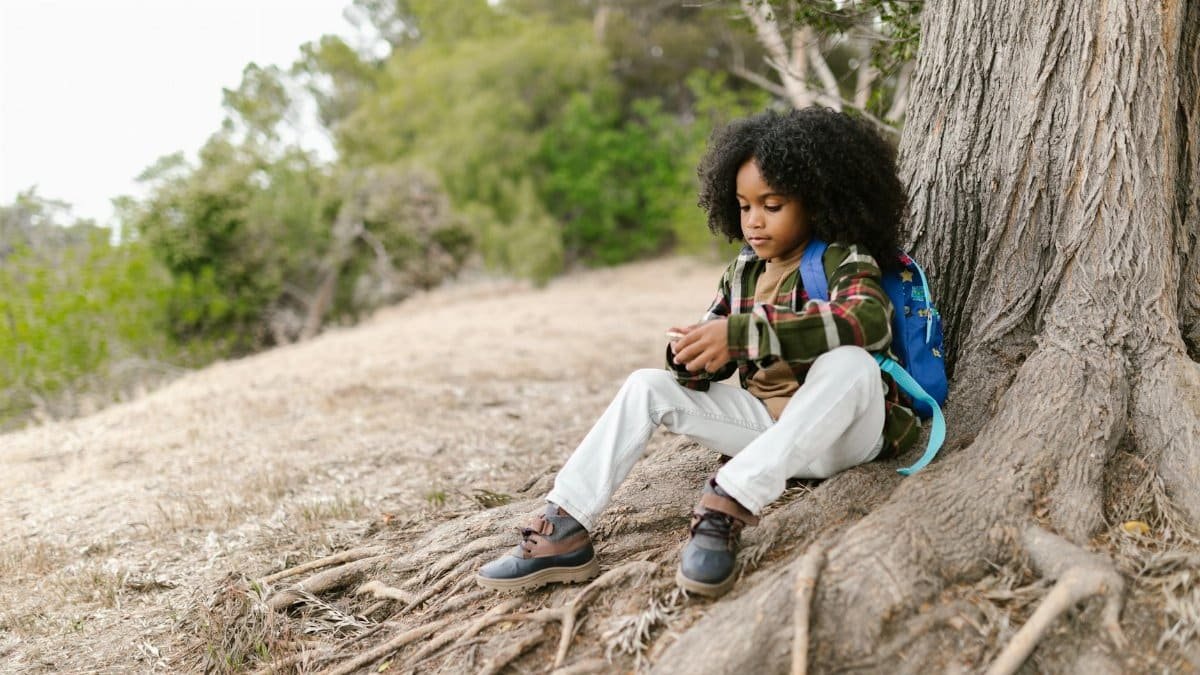 A young boy with curly hair sits against a tree outdoors, wearing a plaid shirt and backpack, enjoying nature.