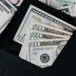 From above of dollar bills in opened black envelope placed on stack of United states cash money as concept of personal income